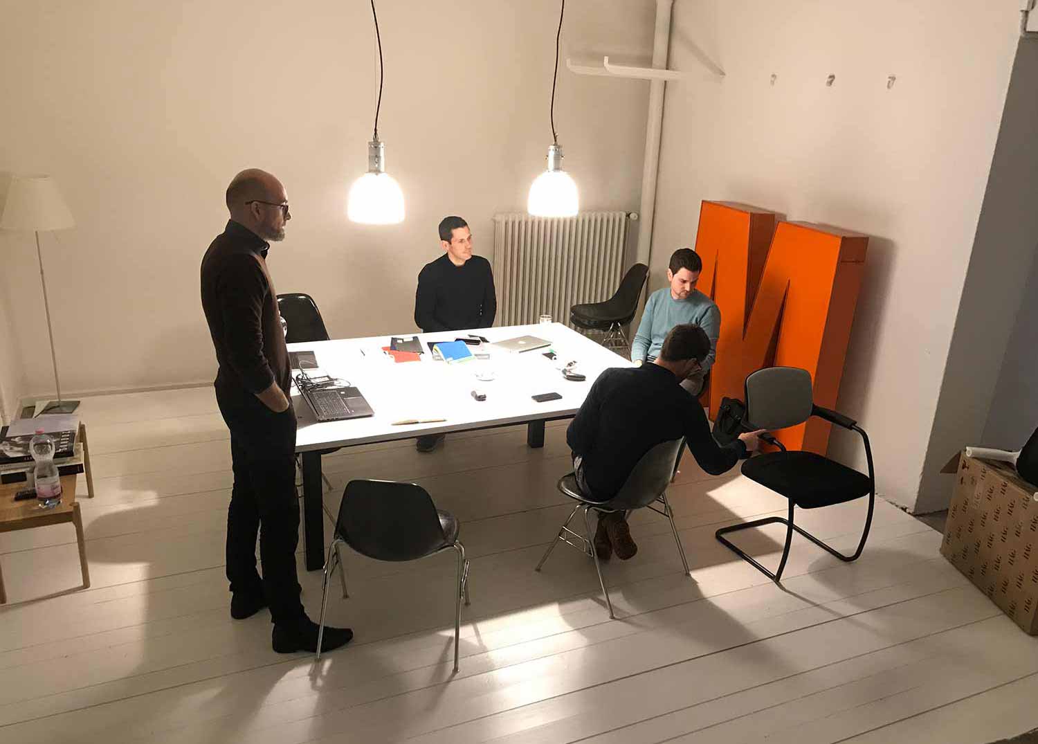 designewrs standing and sitting around a table with 2 pendant lights in a white room